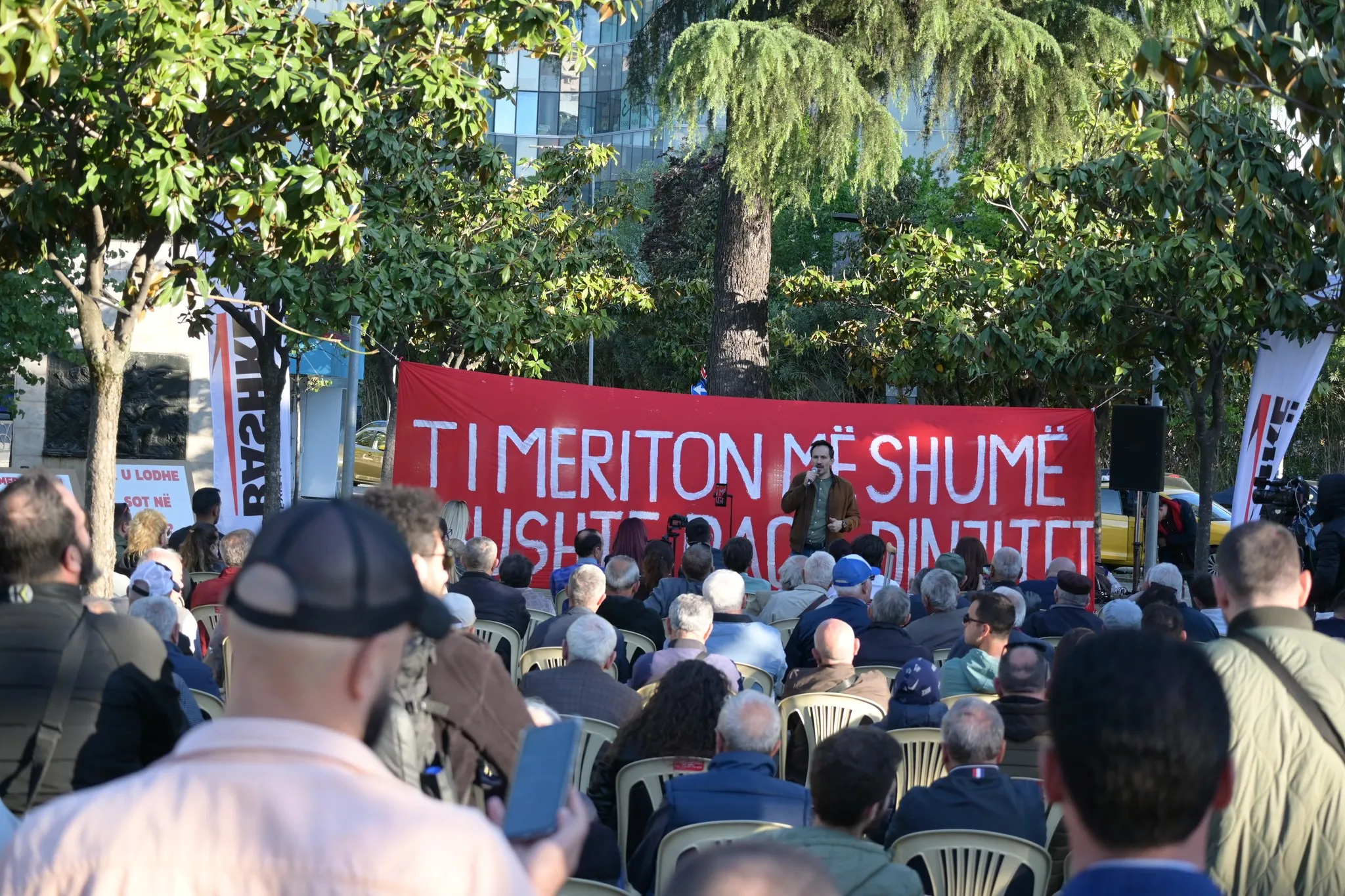 Pamje nga tubimi i për ditën e 1 majit, nje banderle e kuqe shkruan 'Ti meriton më shumë', Redi Muçi duke mbajtur fjalim, dhe pjesëmarrësit të ulur në karrige para tij.