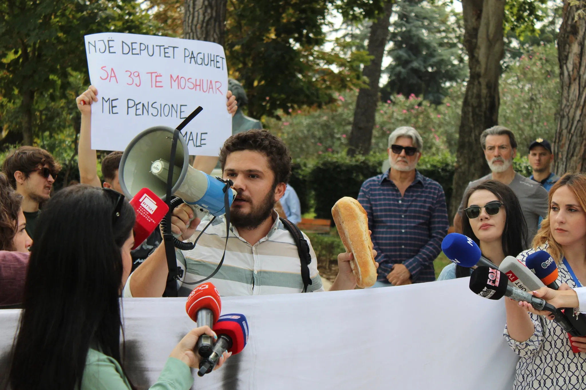 Klodi Leka duke folur ne protesten e Minimumit Jetik me nje buke ne dore, dhe nje pankarte pas tij e cila shkruan: Nje Deputet paguhet sa 39 te moshuar me pension minimal